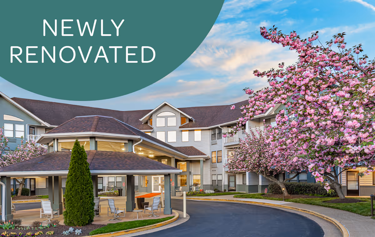 Exterior view of the Virginian senior living facility with a covered entrance, blooming pink cherry blossom trees, and a clear sky. The building is newly renovated as indicated by the text overlay.