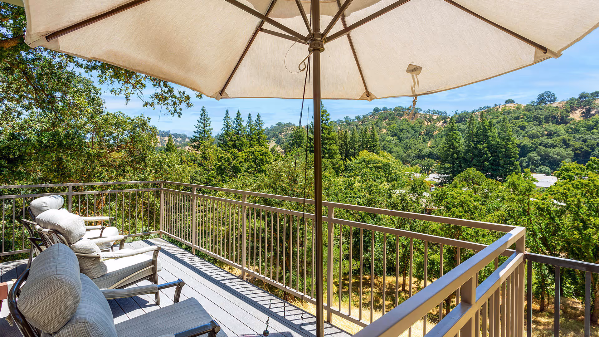 Outdoor balcony with cushioned chairs and a large umbrella overlooking a scenic view of green trees and hills under a clear blue sky.