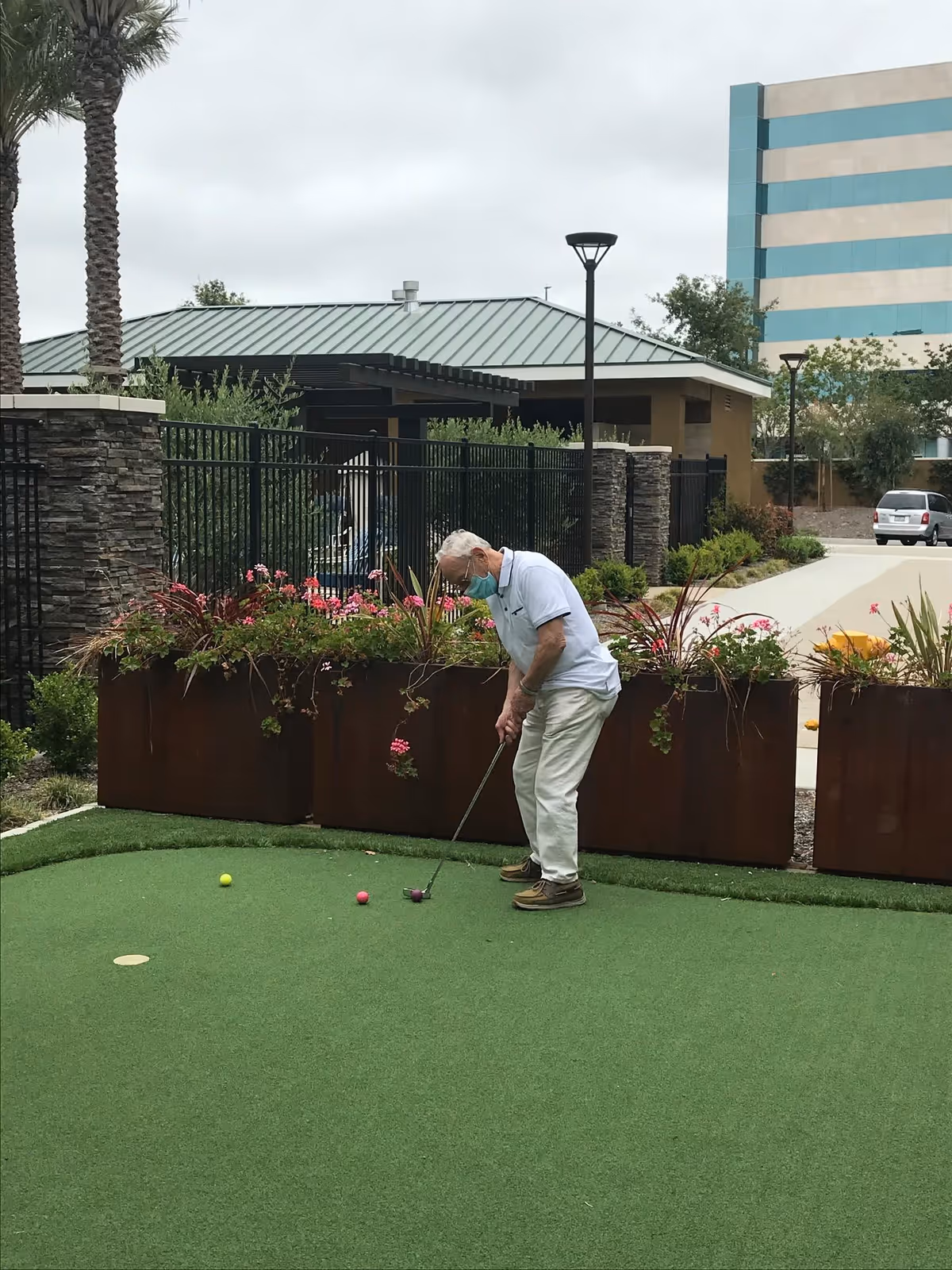 A man wearing a face mask puts a golf ball on a small outdoor artificial putting green next to raised planters and buildings.