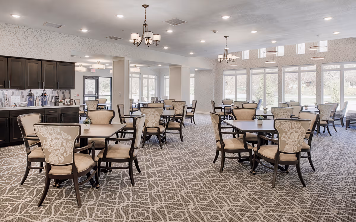 Bright, spacious dining room with multiple round tables, upholstered chairs, large windows, and a service counter.