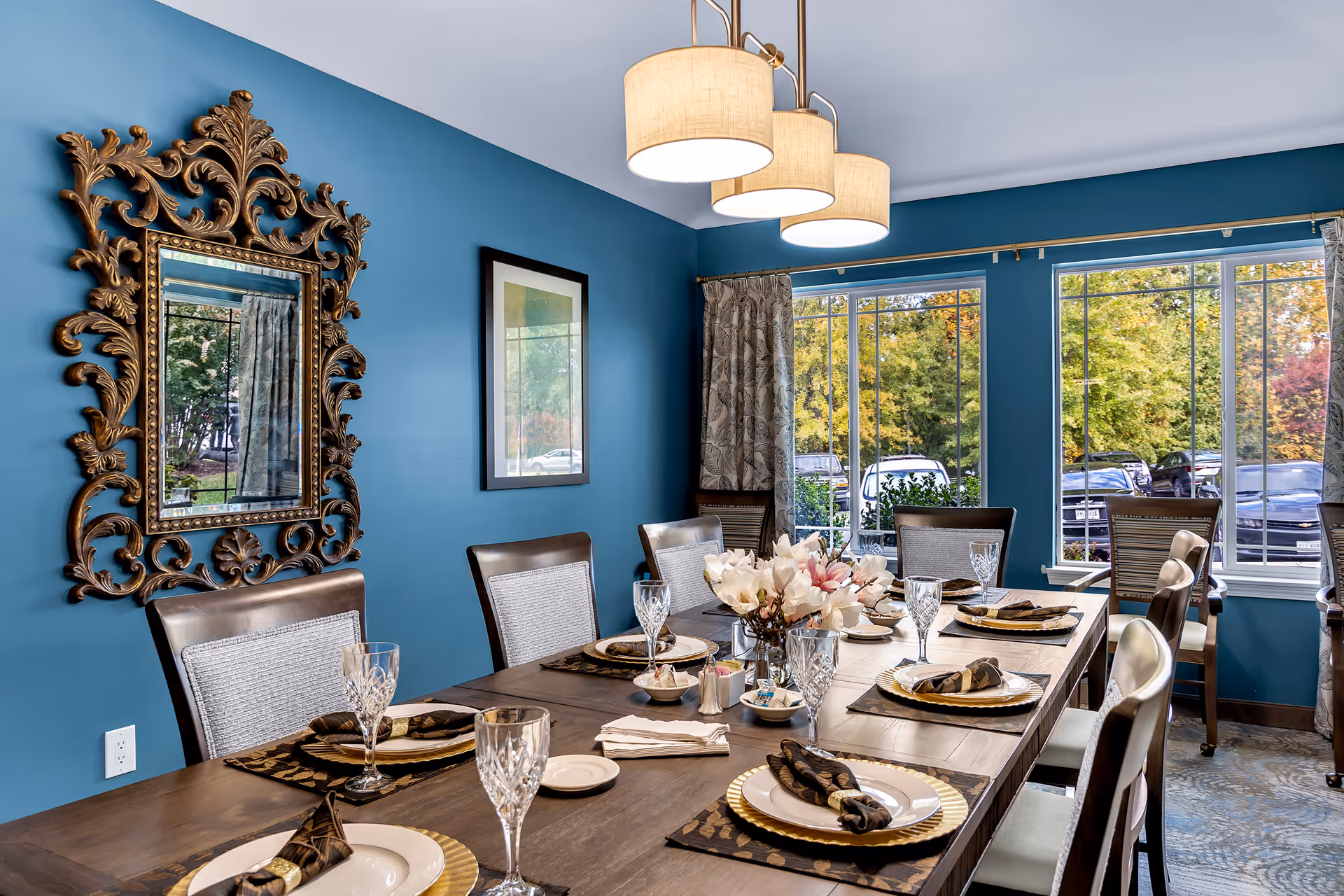 A dining room with a long wooden table set for eight people. Each place setting includes a white plate on a gold charger, a folded dark napkin, and a crystal glass. The room has blue walls, a large ornate gold-framed mirror, a framed picture, and large windows showing trees and parked cars outside. A modern light fixture with three lampshades hangs above the table.
