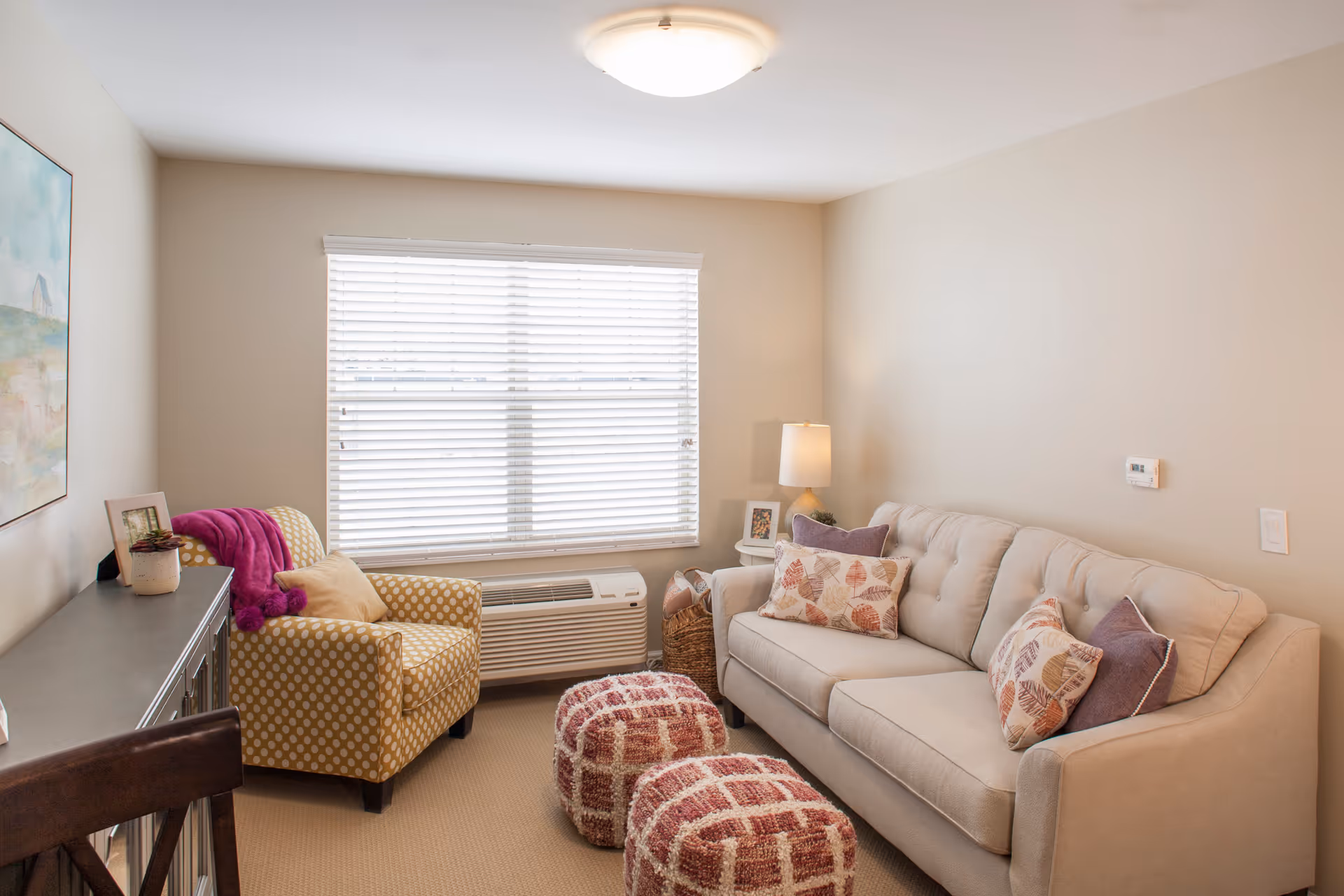 Bright small living room with a beige sofa, patterned armchair, two ottomans, and a window with blinds.