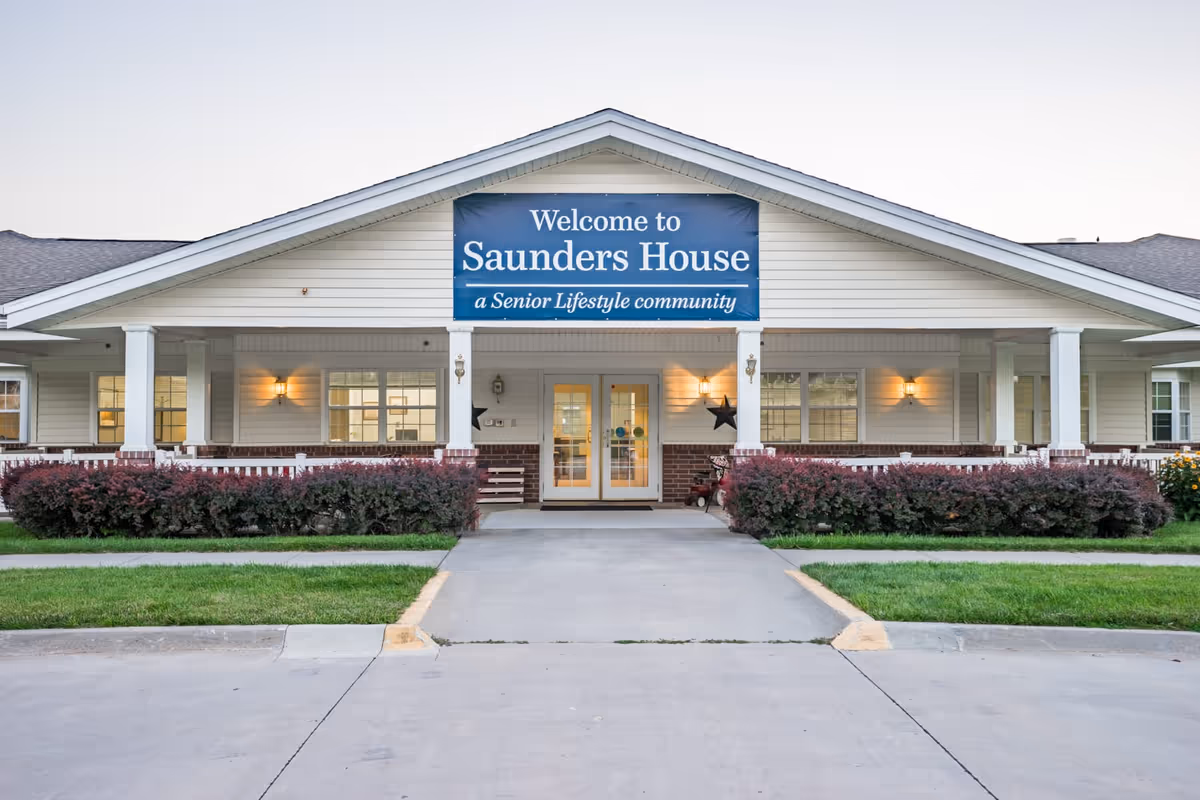 Front exterior view of Saunders House, a senior lifestyle community, showing a welcoming entrance with a large blue sign above the door, white siding, brick accents, and well-maintained landscaping with bushes and grass.