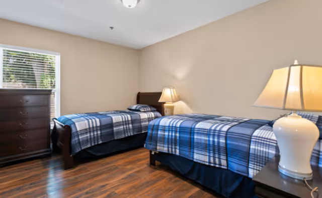 A bedroom with two twin beds dressed in blue plaid bedding, wooden floors, a dresser by a window, and two bedside lamps.