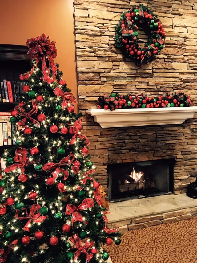 A decorated Christmas tree with red and green ornaments and red plaid bows stands next to a stone fireplace with a fire burning inside. Above the fireplace is a white mantel adorned with a garland of red and green ornaments and plaid ribbons, and a matching wreath hangs on the stone wall above the mantel. A bookshelf with books is partially visible to the left of the tree.