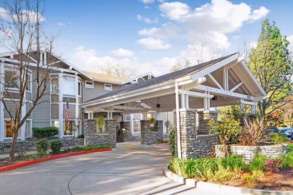 Exterior view of Ivy Park at Walnut Creek senior living facility showing the entrance with a covered driveway supported by stone pillars, surrounded by landscaped greenery and trees under a blue sky with scattered clouds.