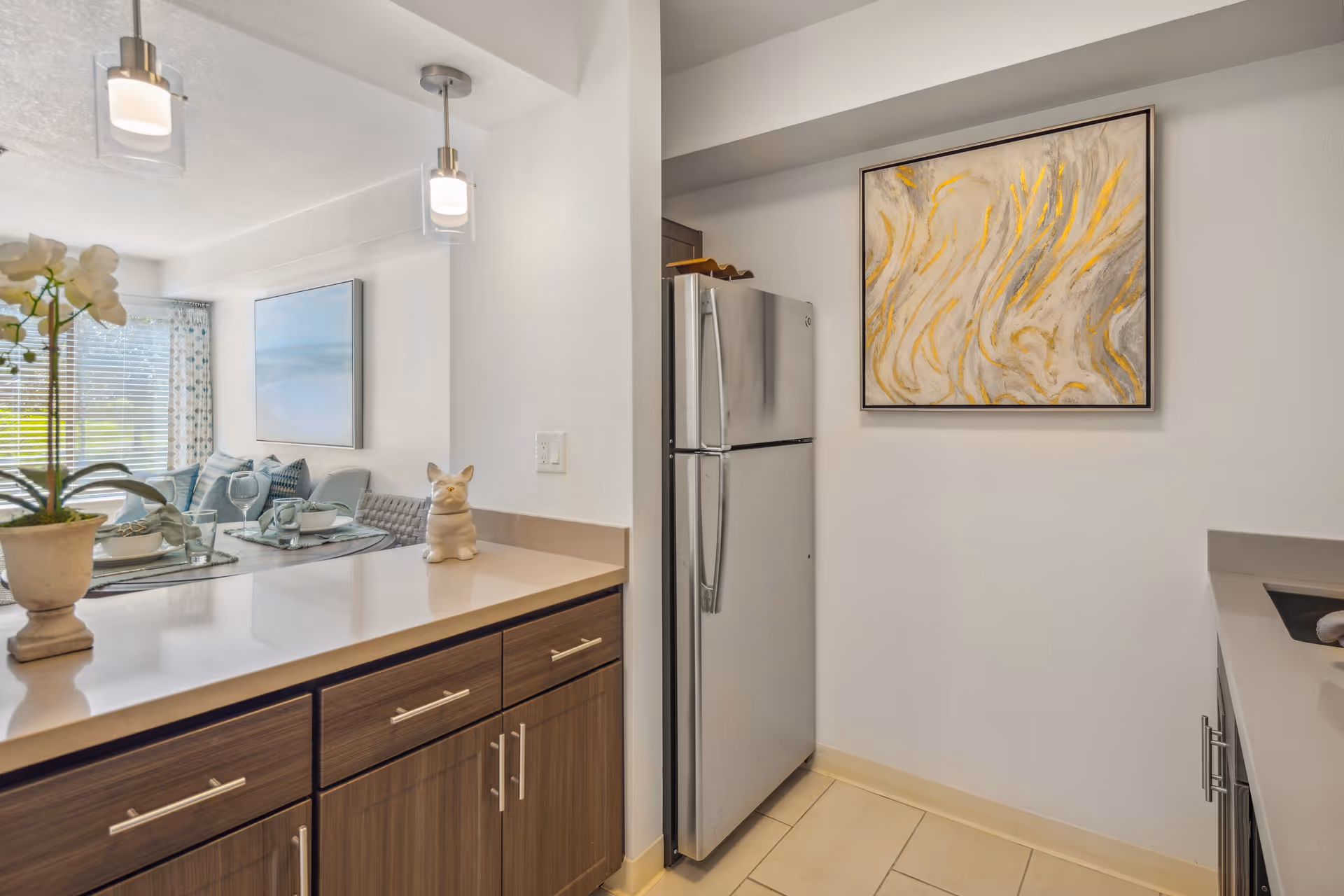 Modern kitchen area with wooden cabinets, a stainless steel refrigerator, and a countertop decorated with a small ceramic dog figurine and a potted plant. In the background, a dining area is visible with a table set for four and a large window with blinds and patterned curtains. Two pendant lights hang from the ceiling, and a framed abstract painting with yellow and gray tones is on the wall near the refrigerator.