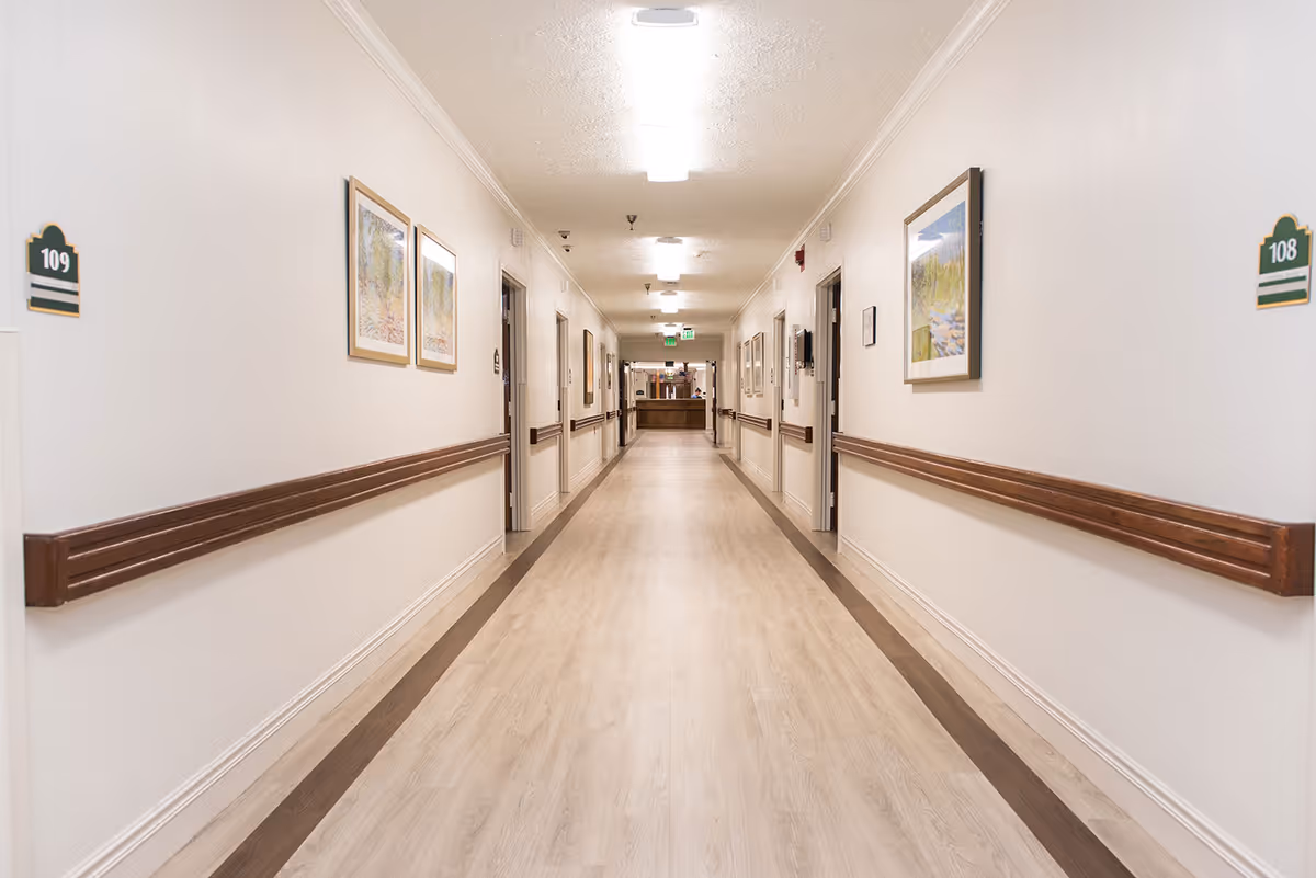 Long, well-lit interior hallway of a senior living facility with handrails, framed artwork, room number signs (108 and 109) and a reception desk at the far end.