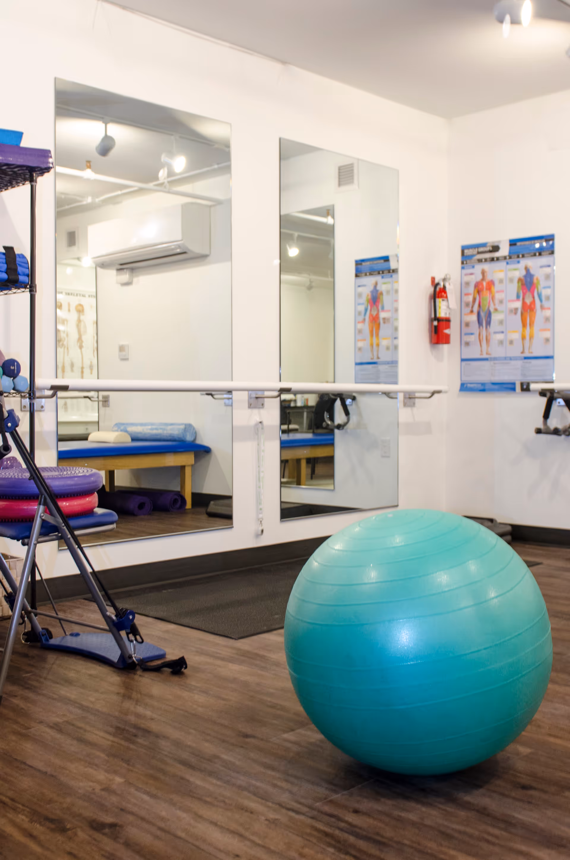 A fitness or physical therapy room with a large turquoise exercise ball on a wooden floor. There are two large mirrors on the wall with a ballet barre in front of them. A rack with various exercise equipment, including weights and balance pads, is visible on the left. Anatomical charts and a fire extinguisher are mounted on the white walls.
