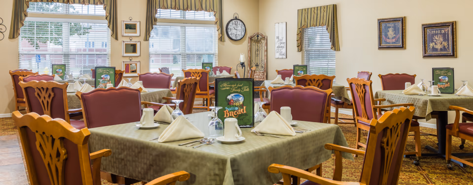 A dining room with multiple tables covered in green tablecloths, each set with white napkins, cups, and silverware. The room has wooden chairs with red cushions, large windows with blinds and valances, and framed artwork on the walls.
