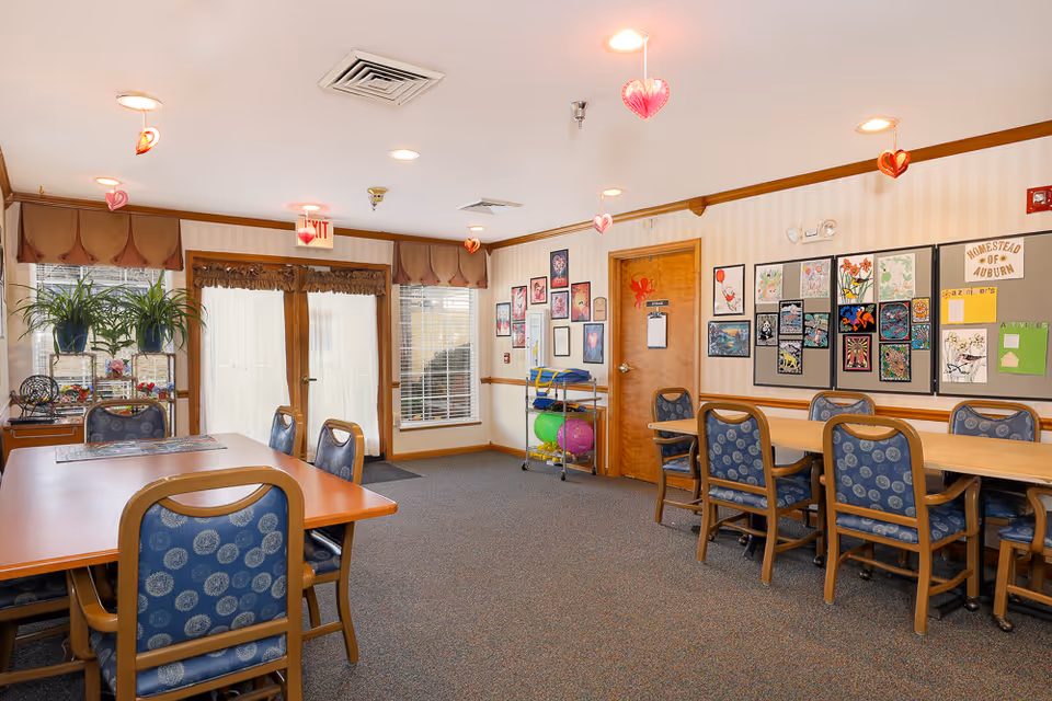 A bright and cozy activity room in an assisted living facility with tables and chairs arranged for group activities. The walls are decorated with colorful artwork and bulletin boards. There are large windows with curtains, potted plants on a shelf, and hanging heart decorations from the ceiling. Exercise balls and equipment are stored on a rack near a wooden door.
