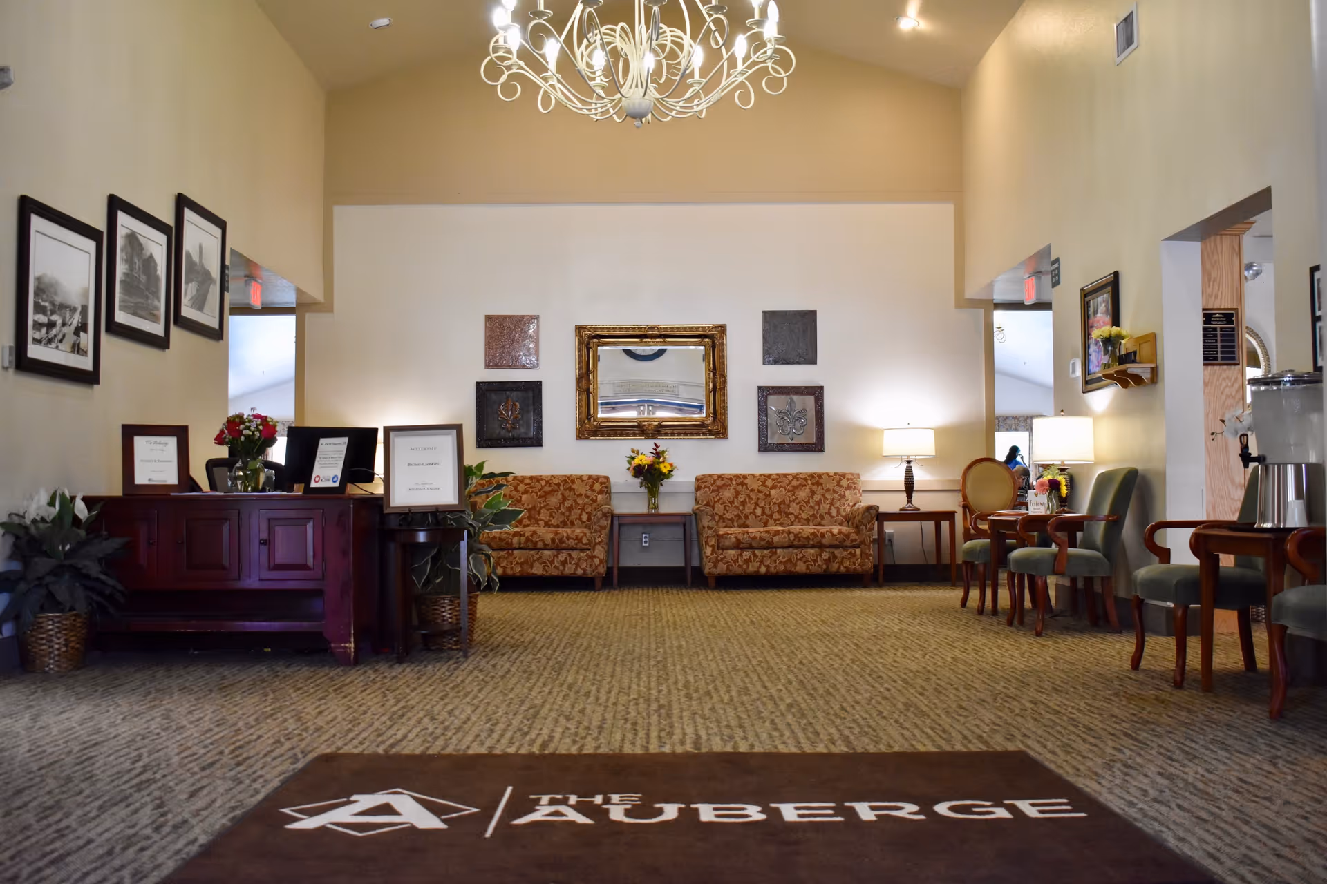 Interior view of a senior living facility lobby with a chandelier hanging from the ceiling, two patterned sofas against the back wall, a mirror and decorative wall art above them, a wooden reception desk on the left with framed signs and flowers, and several chairs with small tables and lamps on the right side. A brown mat on the floor near the entrance displays the logo and text 'THE AUBERGE'.
