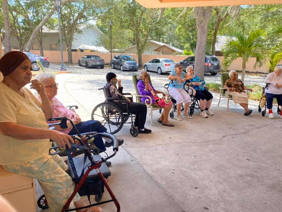 A group of elderly women sitting outdoors under a covered area at Palm Terrace Assisted Living Facility & Adult Day Care Center. Some are seated in wheelchairs and others in chairs, with trees, parked cars, and a fence visible in the background.