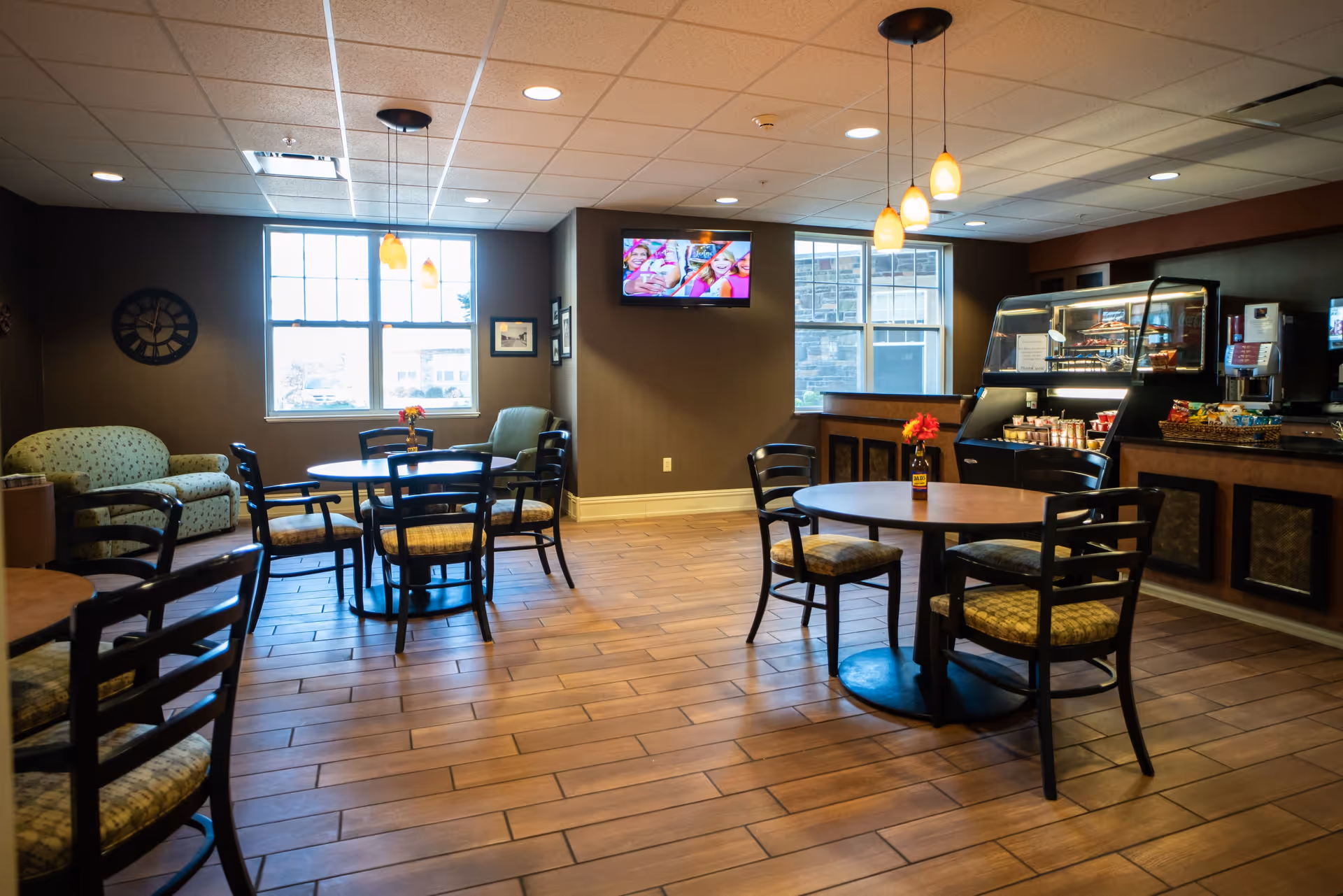 A cozy dining area with round tables and chairs, a small couch, and a display case with snacks and beverages. The room has large windows letting in natural light, a wall-mounted TV, and pendant lights hanging from the ceiling.