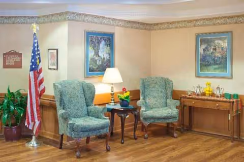 Interior lobby area with two teal armchairs, a lamp, flag, paintings, and a wooden side table.