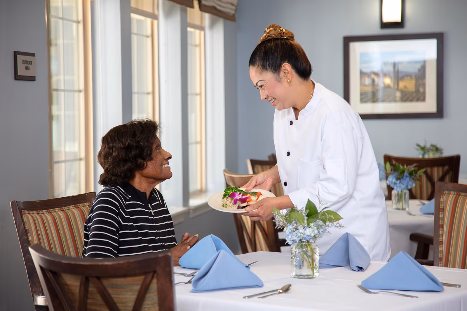 A smiling chef in a white uniform serves a plate of food to a seated elderly woman in a dining room with large windows and tables set with blue napkins and floral centerpieces.