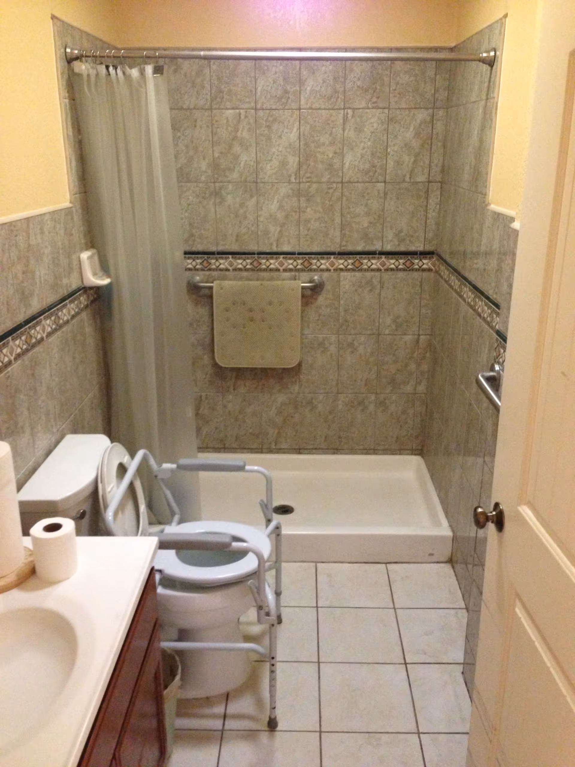 Tiled bathroom with a walk-in shower, wall grab bars, and a raised commode frame over the toilet.