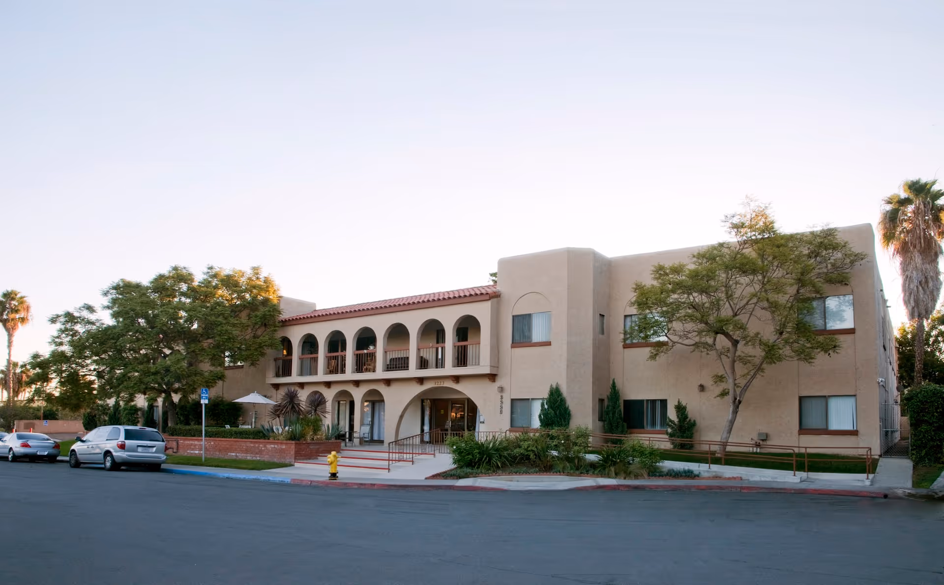 Exterior view of a two-story beige building with arched balconies and a red-tiled roof, surrounded by trees and parked cars along the street in front.