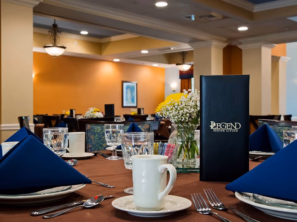 A dining table set with white plates, silverware, glasses, a white coffee cup, and blue folded napkins in a senior living facility dining room. A vase with yellow and white flowers and a black menu with the text 'Legend Senior Living' are also on the table. The background shows more tables and chairs with warm lighting and orange walls.