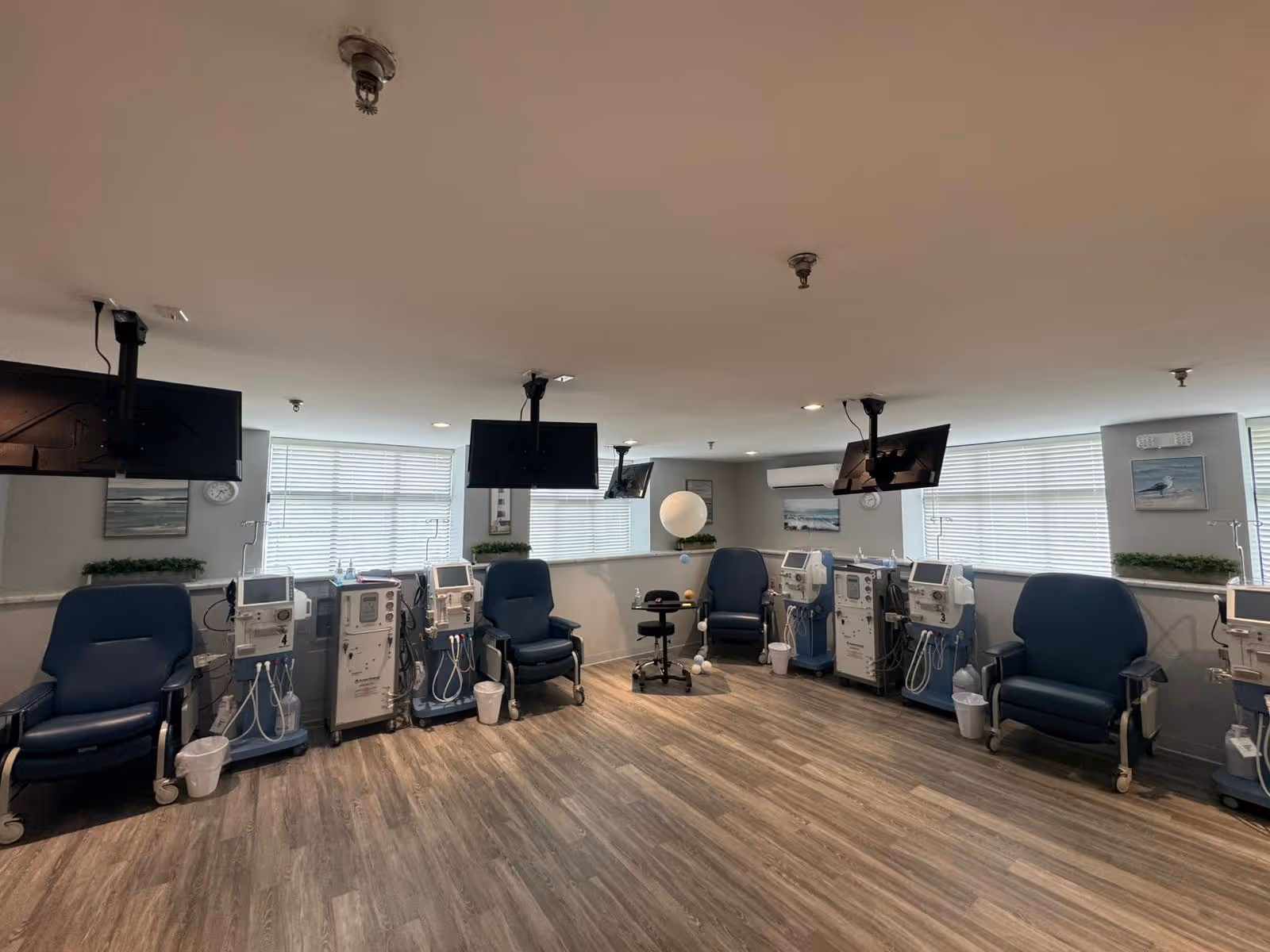 A clinical treatment room with multiple reclining chairs, dialysis machines along the walls, and ceiling-mounted TVs.