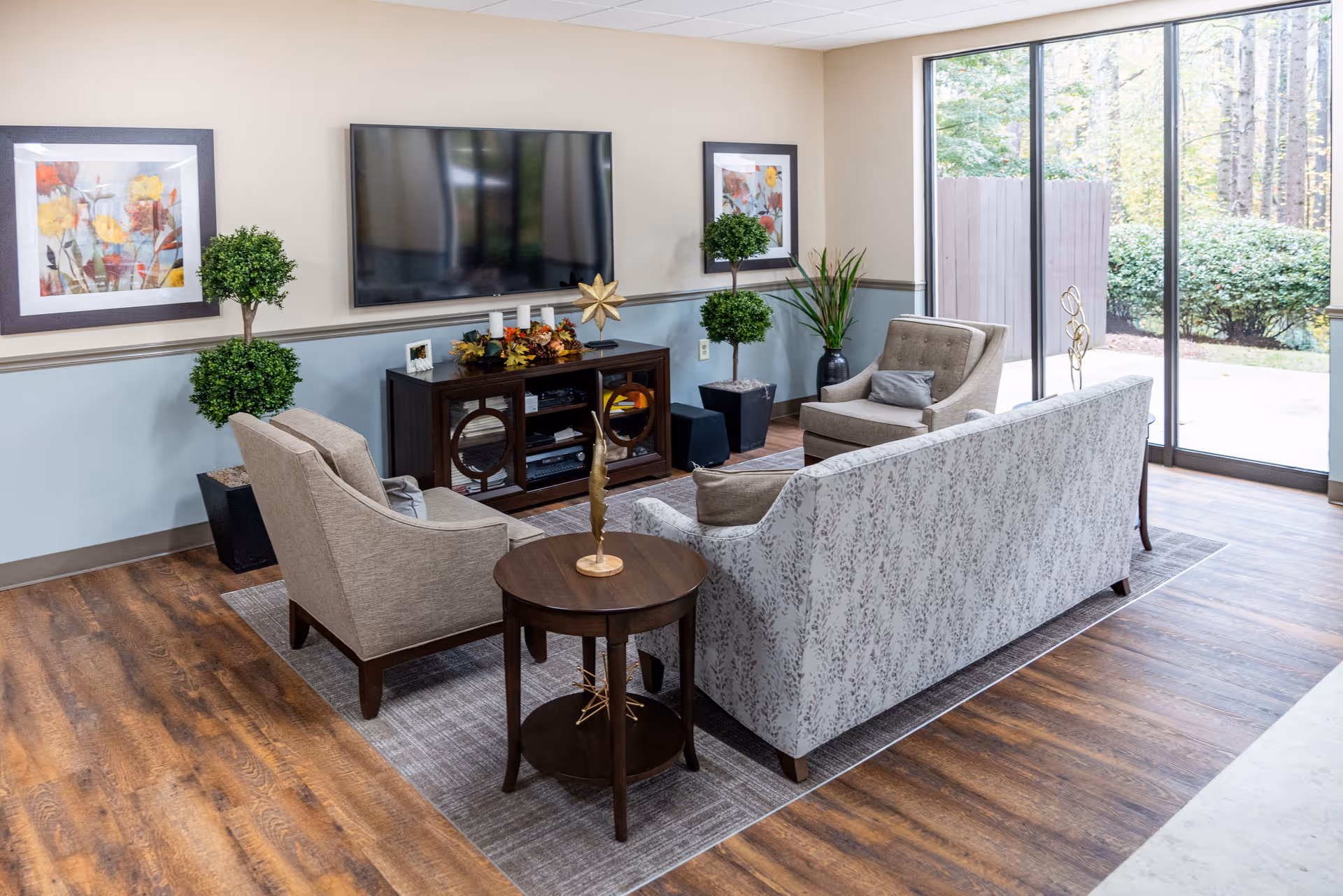A cozy senior living room with a patterned sofa, two armchairs, a round wooden side table, and a TV mounted on the wall. The room has wooden flooring, two framed floral paintings, potted plants, and large glass sliding doors that open to an outdoor area with greenery.