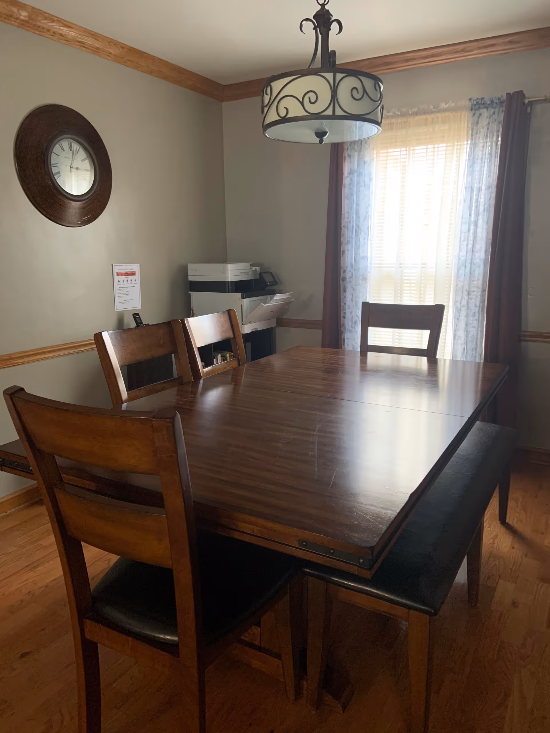 A dining room with a wooden table surrounded by four wooden chairs and a bench. A large window with sheer and dark curtains allows natural light to enter the room. A decorative wall clock hangs on the left wall, and a printer is placed on a small table in the corner. A ceiling light fixture with a decorative metal frame hangs above the table.