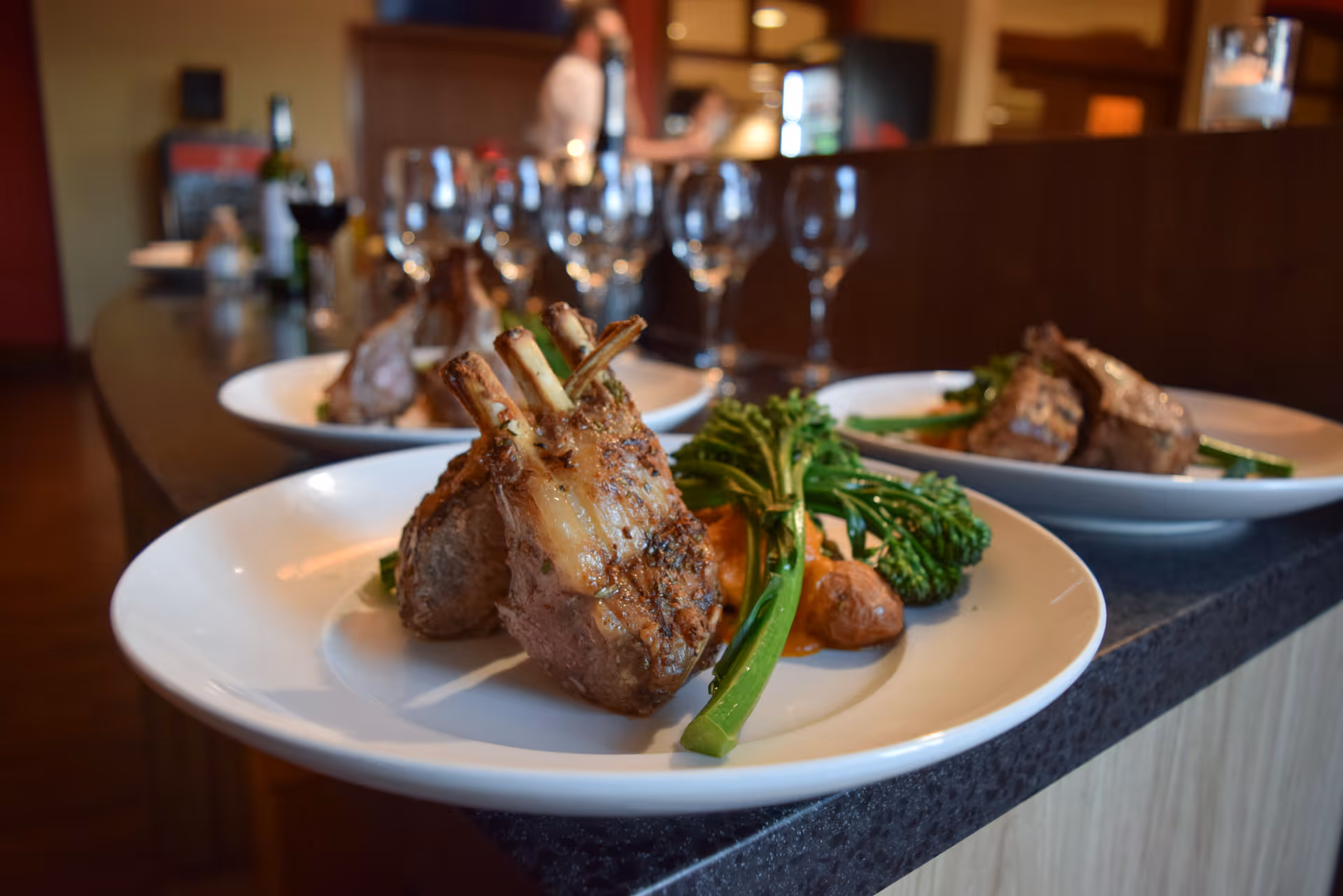 Close-up view of plated gourmet meals featuring grilled lamb chops, roasted potatoes, and steamed broccolini on white plates placed on a dark countertop with wine glasses and a blurred background of a person and kitchen area.