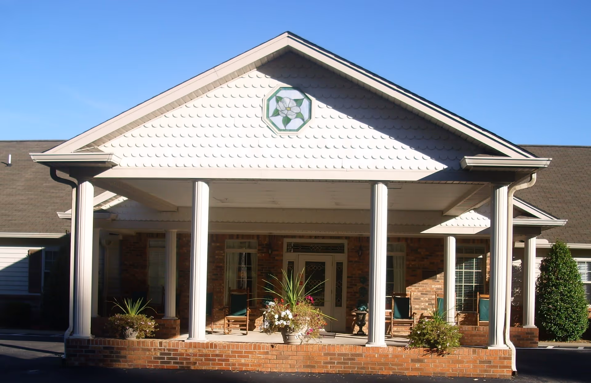 Front exterior view of Magnolia Place Assisted Living building featuring a covered entrance with white columns, brick base, and a decorative emblem on the gable. There are plants and chairs on the porch area under a clear blue sky.