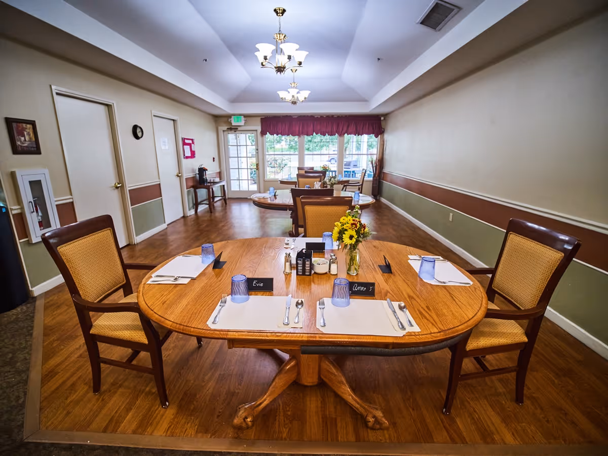 A dining room in a senior living facility with a wooden table set for a meal, chairs, place settings and a vase of flowers.