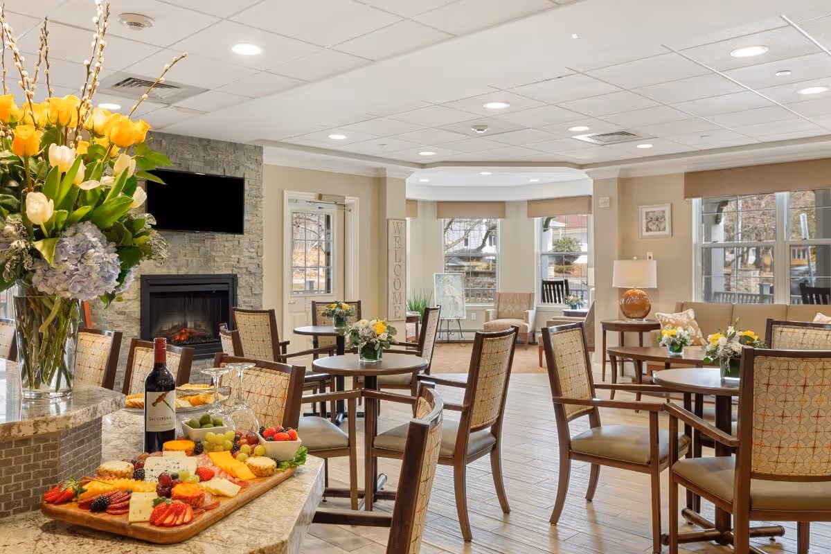 A bright and inviting common area with round tables and cushioned chairs arranged for socializing. A stone fireplace with a mounted TV above it is visible on one wall. A countertop in the foreground holds a large vase of flowers, a bottle of wine, wine glasses, and a wooden board with an assortment of cheeses, fruits, and crackers. Large windows let in natural light, and there are additional seating areas with armchairs and side tables decorated with flowers and lamps.