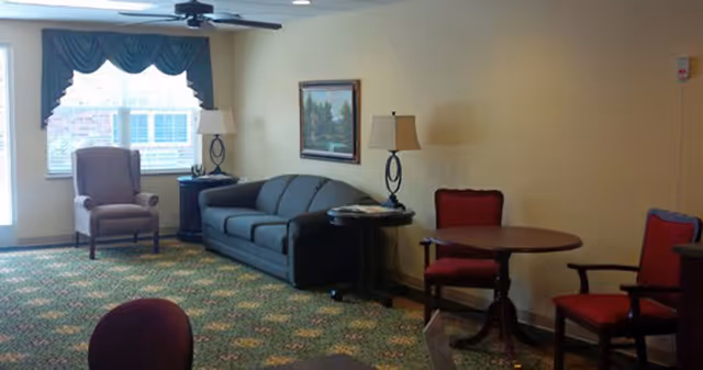 A sitting area in a senior living facility with a dark gray sofa, a purple armchair near a window with green curtains, two table lamps on side tables, a round wooden table with two red cushioned chairs, and a green patterned carpet.