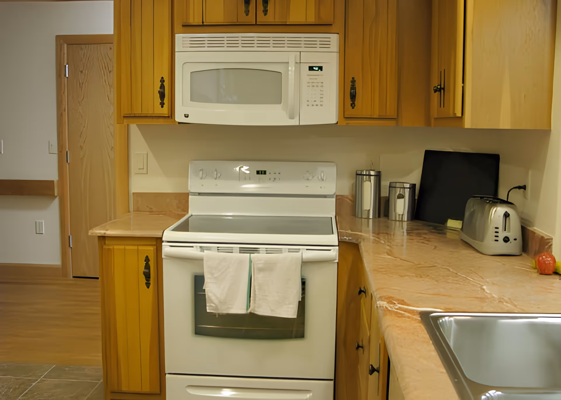 A kitchen area featuring wooden cabinets, a white electric stove with two towels hanging on the handle, a white microwave mounted above the stove, a countertop with a toaster, two canisters, a cutting board, and a sink in the foreground.