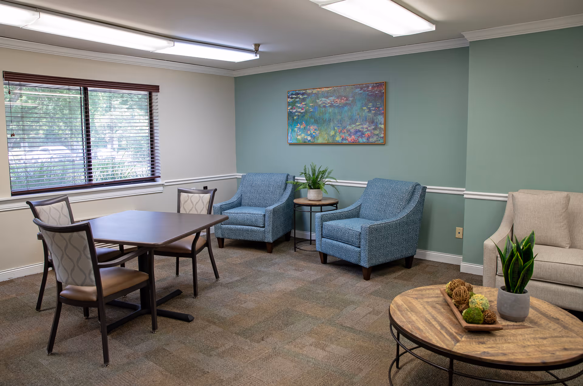 A cozy sitting area in a nursing and rehabilitation center featuring two blue armchairs with a small round table and plant between them, a beige sofa, a wooden coffee table with decorative items and a plant, and a rectangular table with four chairs near a window with blinds. A colorful painting hangs on the green accent wall.