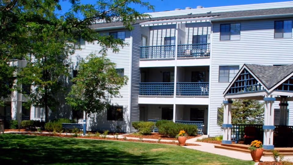 Exterior view of a multi-story residential building with white siding and blue railings, surrounded by green trees, shrubs, and a well-maintained lawn. There is a gazebo with a shingled roof and potted plants near a paved walkway in the foreground.