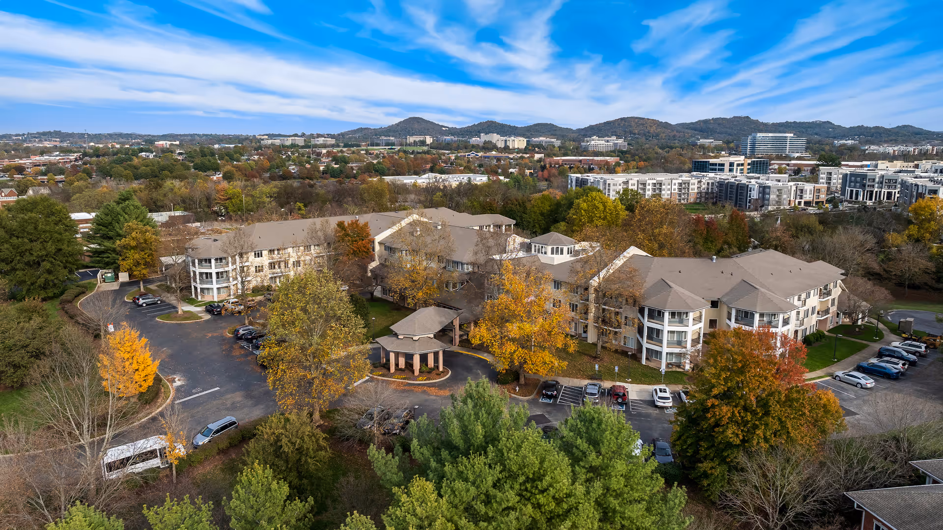 Aerial view of a senior living community building surrounded by trees with autumn foliage, parking lots with cars, and a gazebo at the entrance. In the background, there are hills and a cityscape under a partly cloudy blue sky.