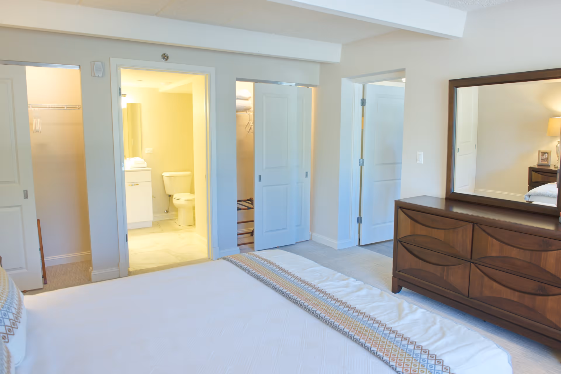 A bright bedroom with a white bedspread featuring a patterned strip, a wooden dresser with a large mirror, and an open doorway leading to a bathroom with a toilet and sink. There are also two closets with sliding doors visible.