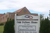 White sign reading 'Odd Fellows Home' in front of a multi-story brick building under a partly cloudy sky.