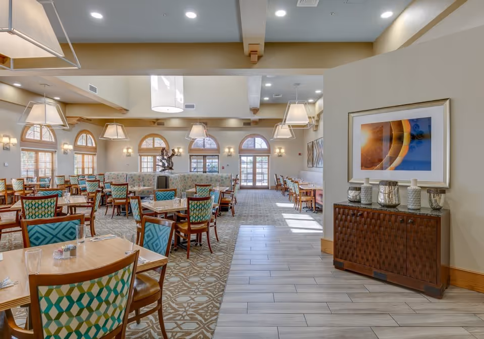 Spacious dining room with multiple wooden tables and chairs featuring patterned upholstery. The room has large arched windows allowing natural light to fill the space. Modern pendant lights hang from the ceiling. A sideboard with decorative vases and a framed abstract artwork is visible on the right side.