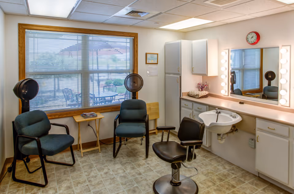Small hair salon room with styling chairs, hooded dryers, a sink, lighted mirror and a large window looking out to a patio.