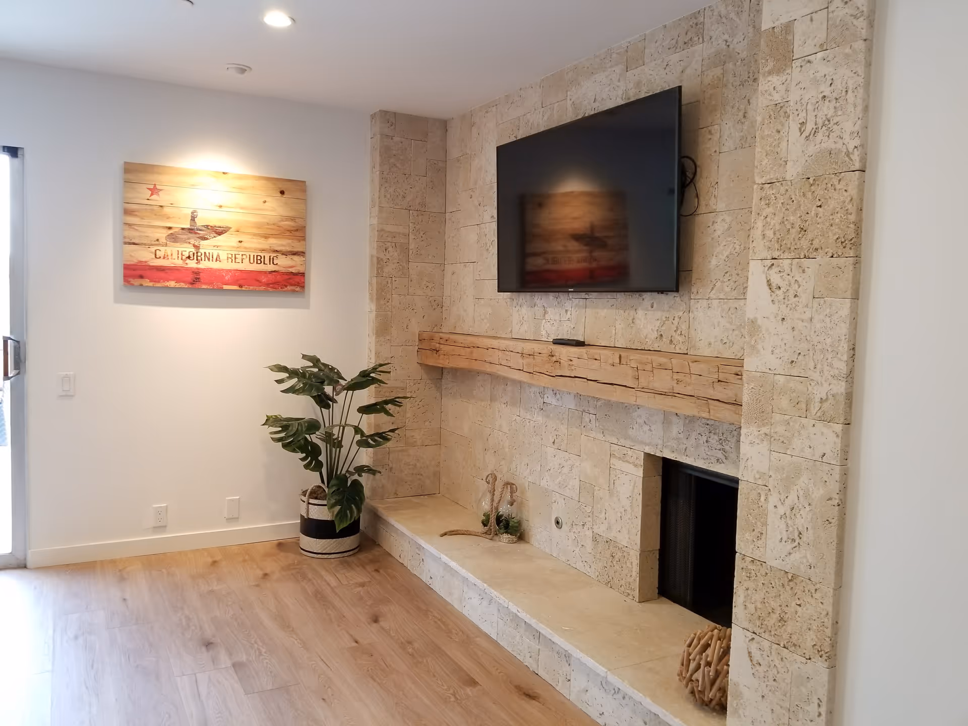 A cozy living room area featuring a stone fireplace with a wooden mantel, a flat-screen TV mounted above it, a potted plant on the floor, and a wooden wall art piece depicting the California Republic flag illuminated by a spotlight.