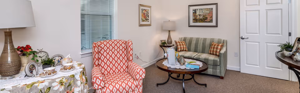 A cozy living room with a patterned red armchair, a green striped sofa with two plaid pillows, a round wooden coffee table with decorative items, a side table with a lamp, and framed artwork on the walls. There is a window with blinds and a white door in the background.
