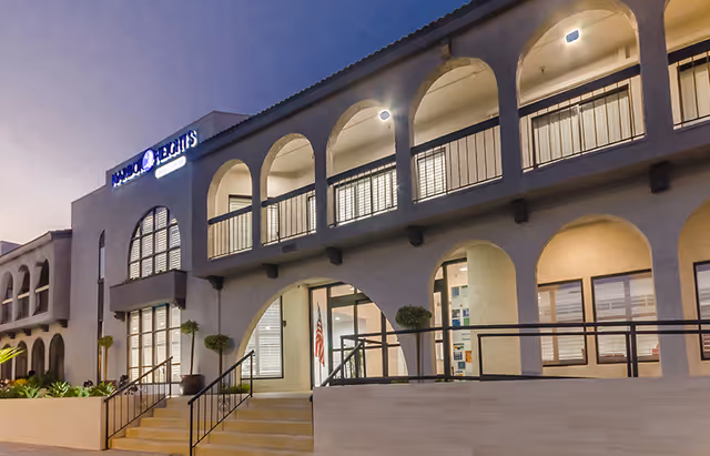 Exterior view of Harbor Heights building at dusk, featuring arched windows and balconies with lights on inside. There are stairs and a ramp leading to the entrance, with plants along the front.