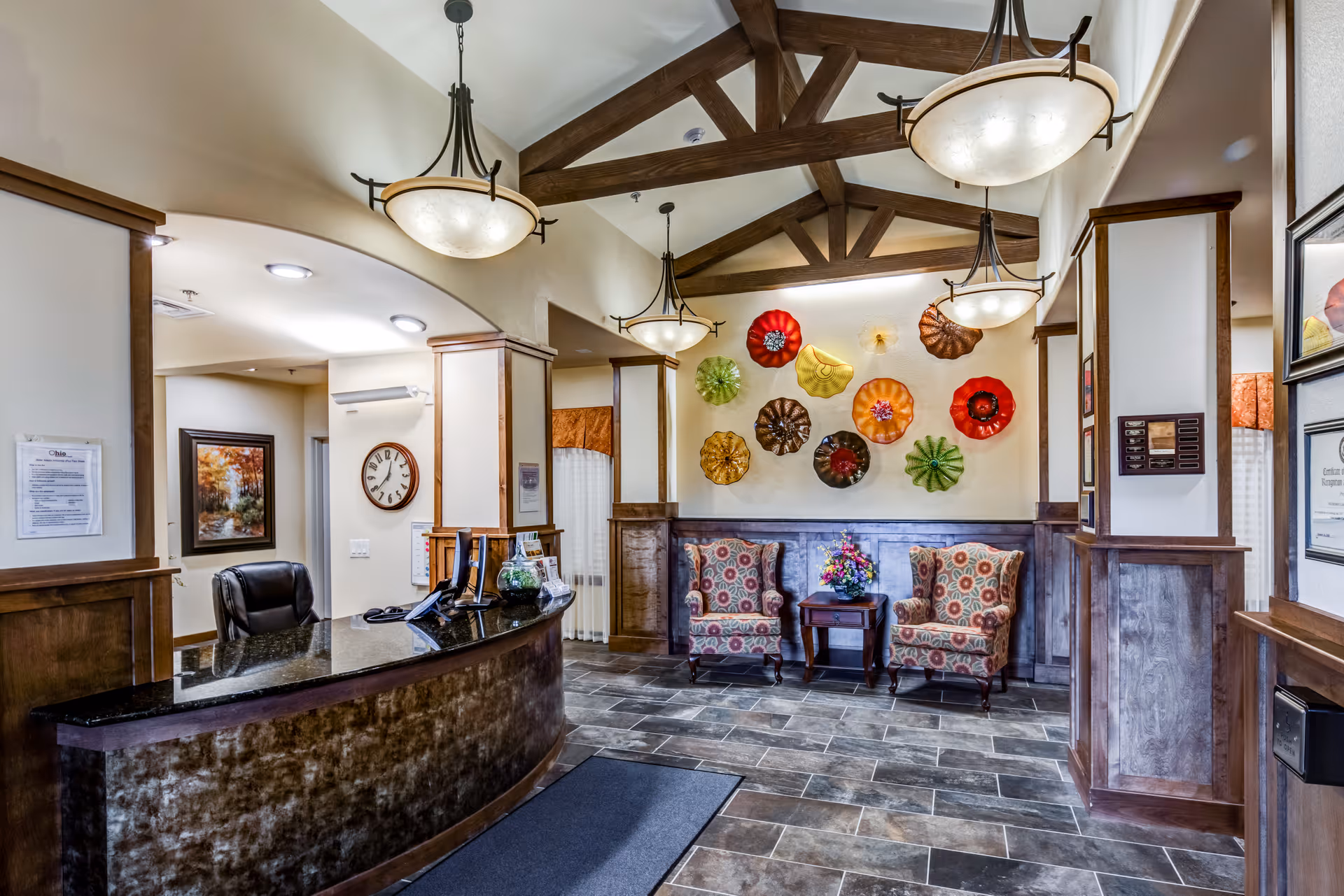 Reception lobby of a memory care facility with a curved front desk, hanging chandeliers, decorative wall art and two patterned armchairs beneath exposed wooden beams.