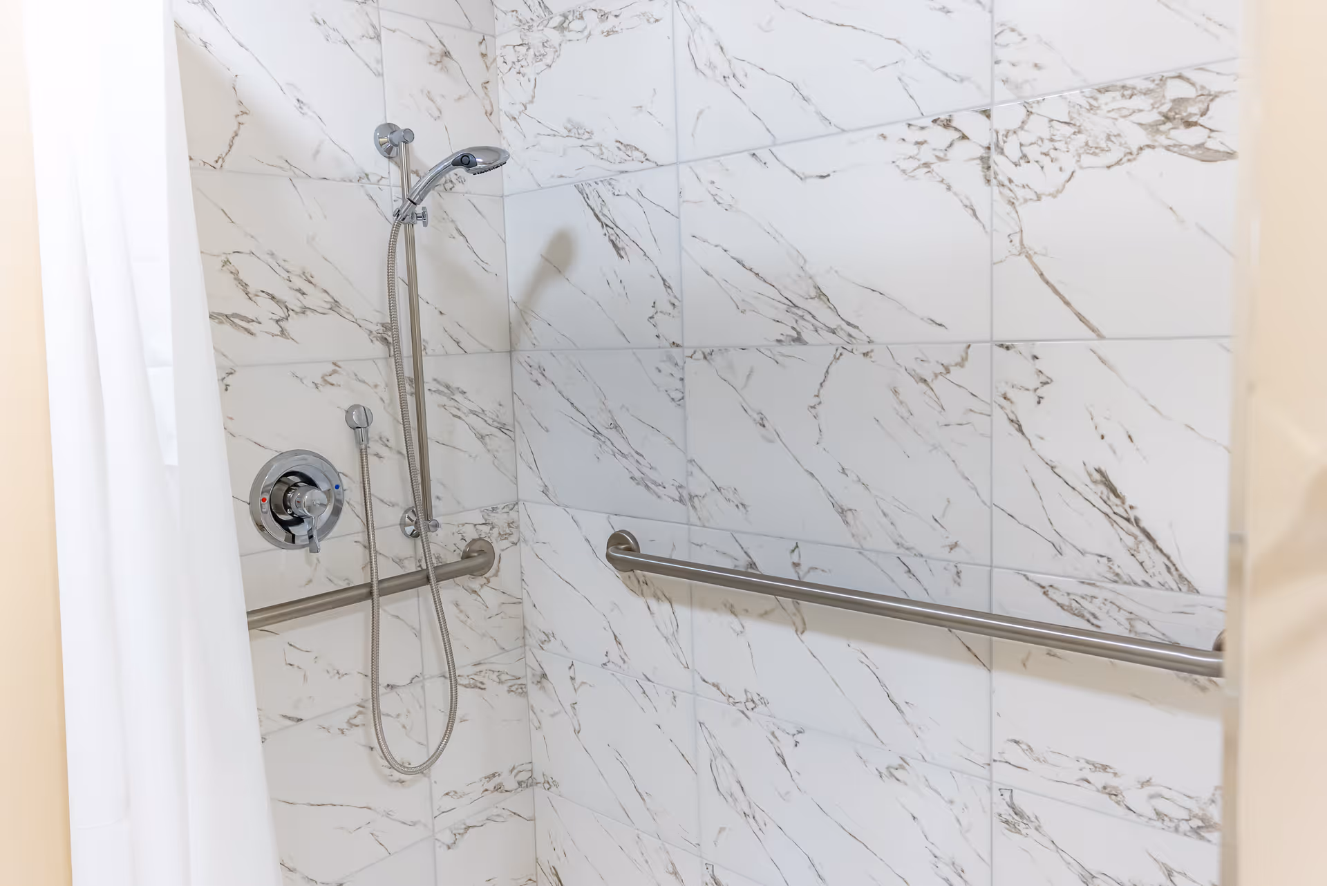 Shower area with white marble-patterned tiles, a handheld showerhead attached to the wall, a control knob, and a horizontal grab bar for support. A white shower curtain is partially visible on the left side.