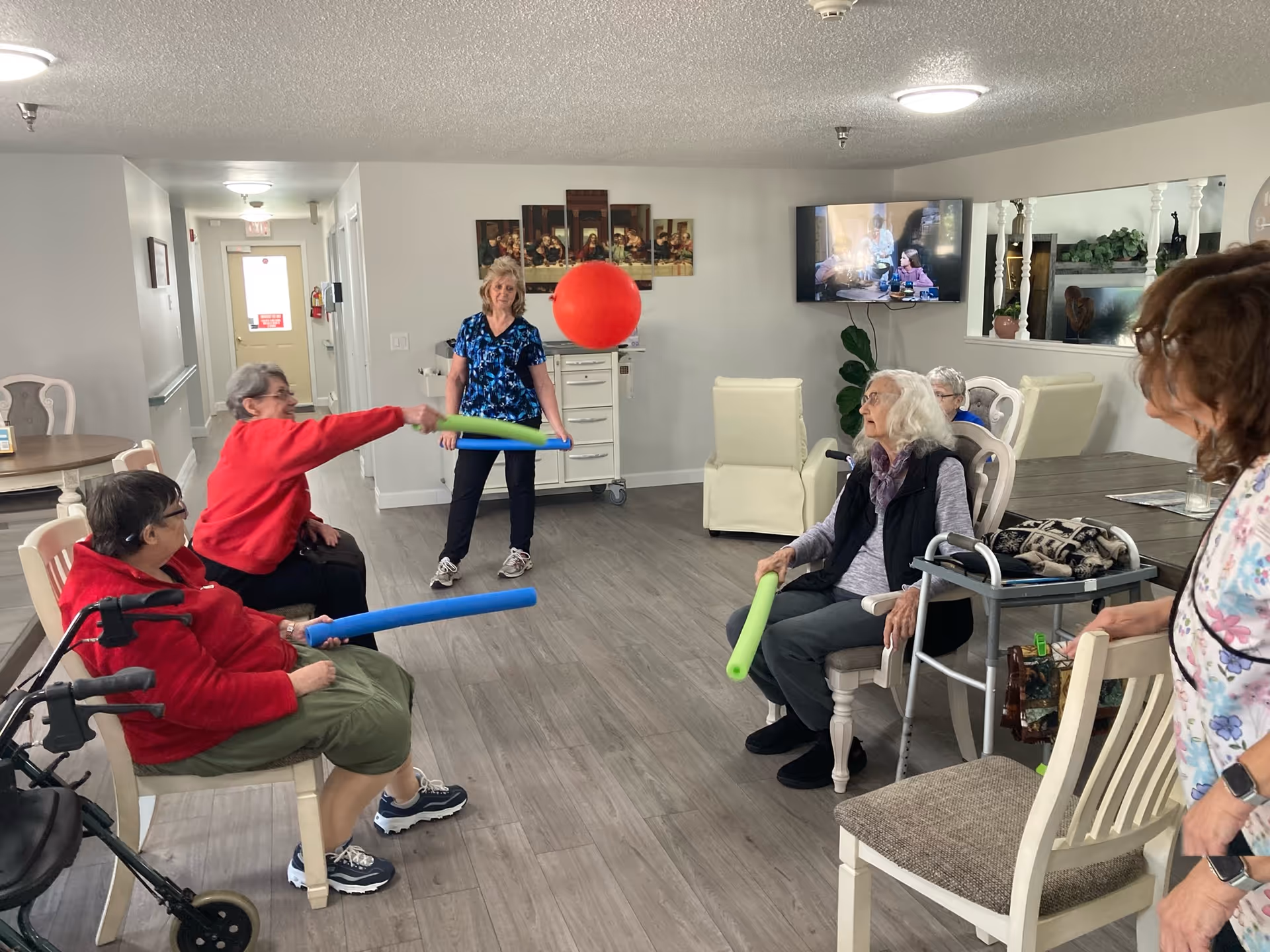 Residents in a communal living room playing a group game with a large red balloon and foam noodles.