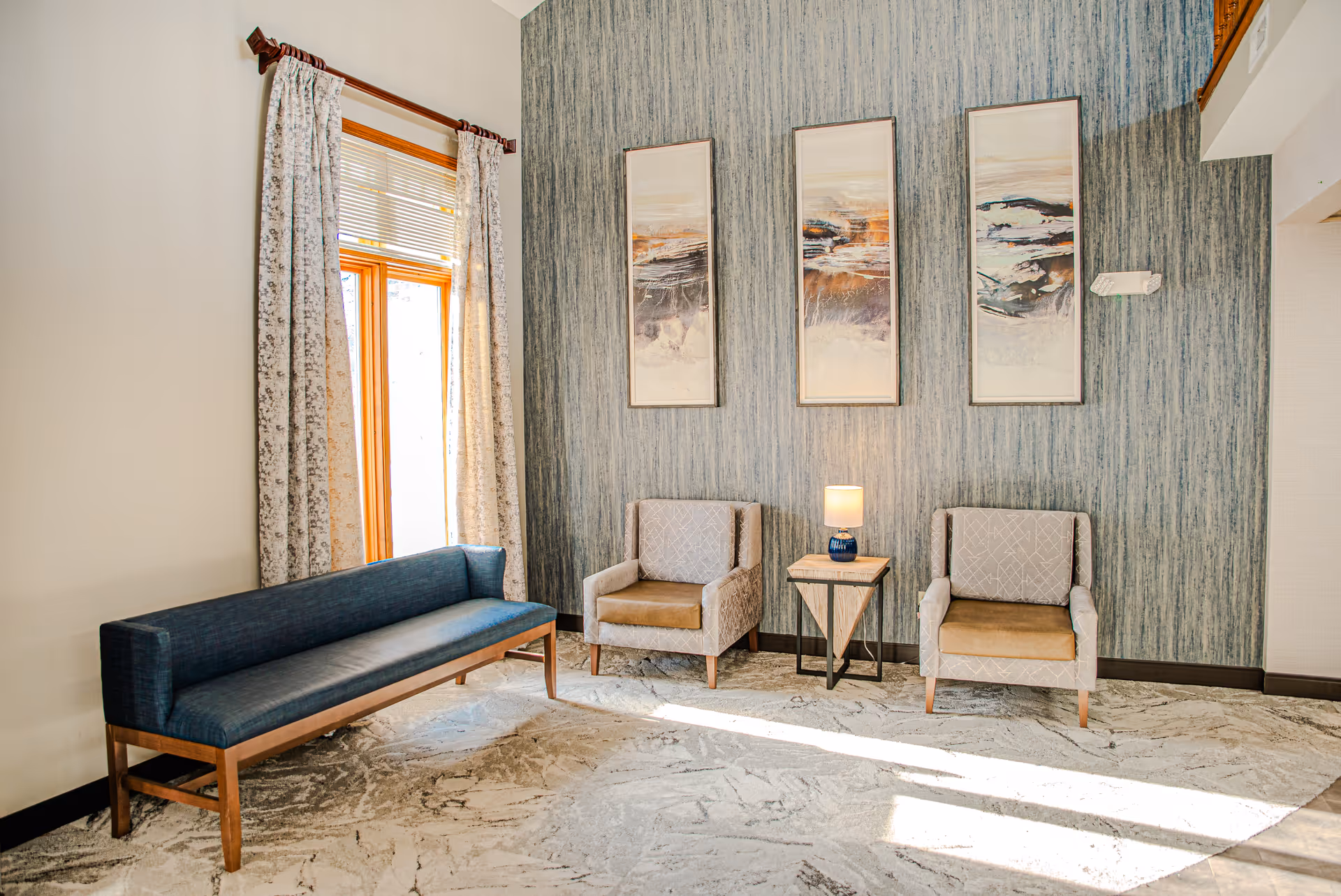 A bright sitting area with a long blue bench on the left, two beige armchairs separated by a small wooden side table with a lamp, and three abstract paintings hanging on a textured blue-gray wall. A window with patterned curtains allows natural light to fill the space.