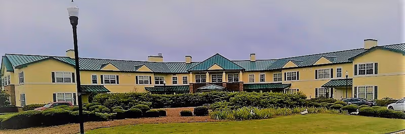 Front exterior view of a large two-story yellow building with green roof, multiple windows, and a landscaped garden with bushes and grass in front. Several cars are parked on the sides and a streetlamp is visible on the left side of the image.