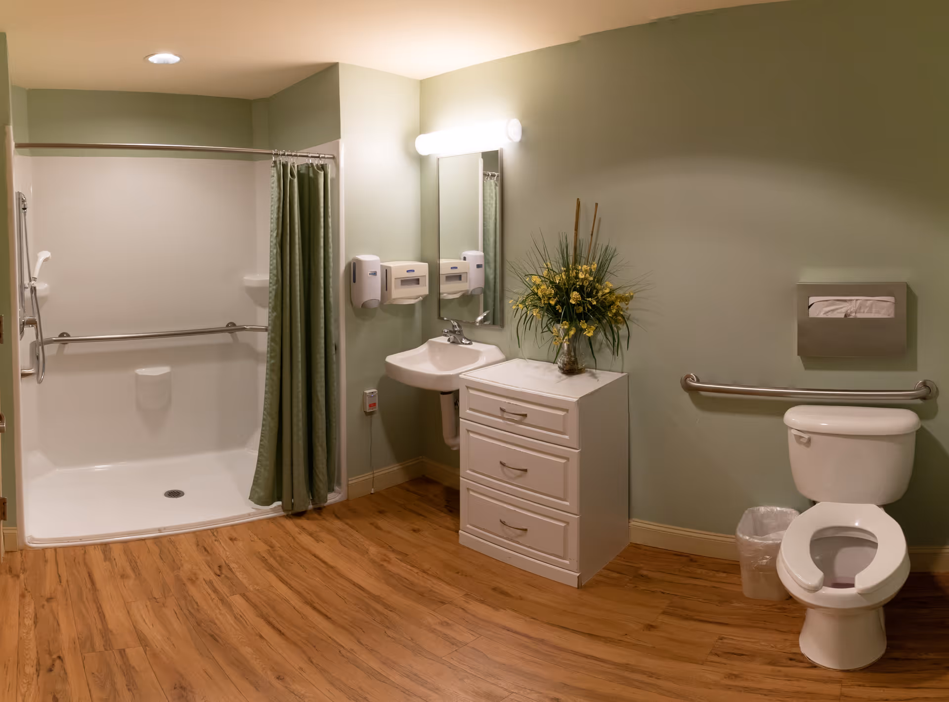 A clean and accessible bathroom with a walk-in shower featuring a green curtain and grab bars, a white toilet with a grab bar beside it, a small white sink with a mirror above, two wall-mounted soap dispensers, a white cabinet with drawers topped with a vase of yellow flowers, and wood-look flooring.