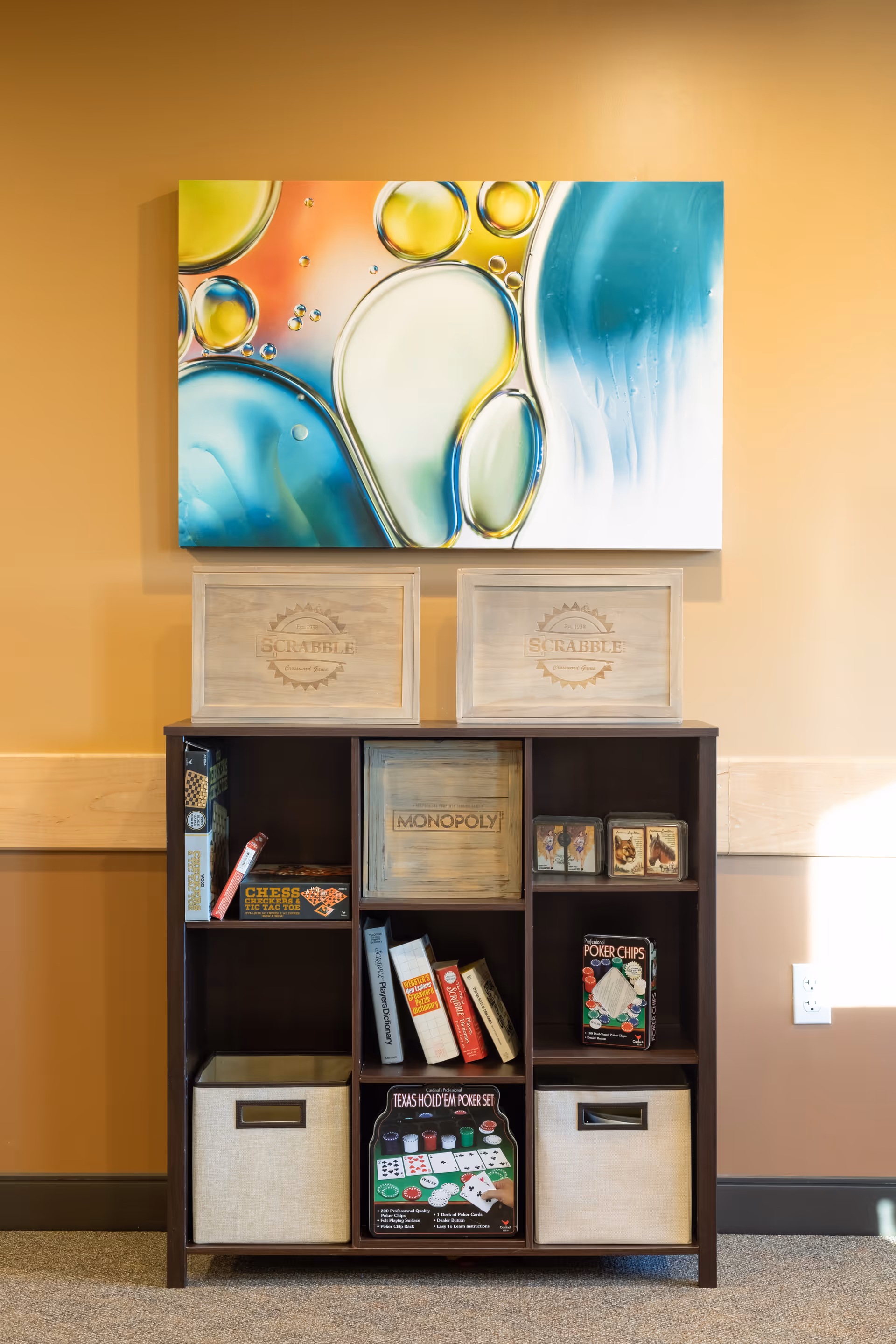 Wooden cubby shelf stocked with board games and fabric bins beneath a colorful abstract painting on a warm-toned wall.