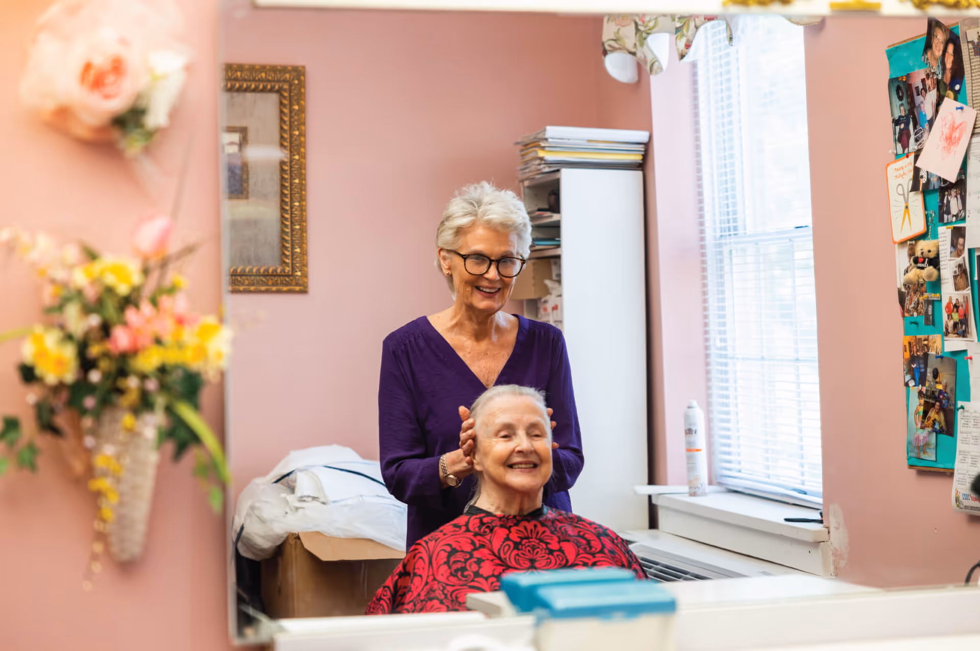 An elderly woman with short white hair and glasses stands behind another elderly woman who is seated and smiling, wearing a red and black patterned cape, in a room with pink walls and a window with blinds. The standing woman appears to be styling or massaging the seated woman's hair. The room has a bulletin board with photos and notes, a floral wall decoration, and a cabinet with stacked papers.